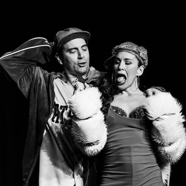 Two performers on stage, with the man wearing a baseball cap and costume reminiscent of wildlife conservation and the woman in a glamorous outfit with a feather boa, both engaging in a dramatic scene inspired by animal behavior