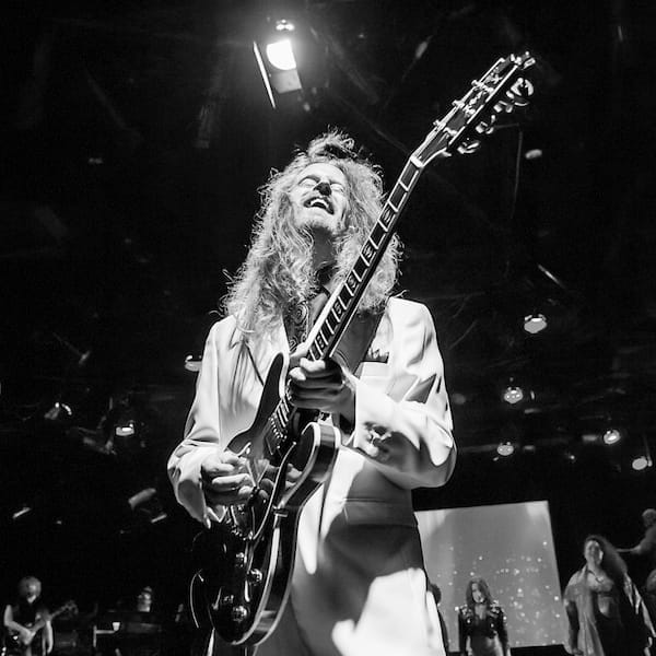 Guitarist in white suit performing on stage in a black and white photo for Evolutionaries Remastered.