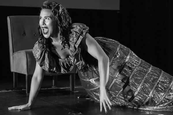 A woman in an elaborate costume with a large skirt performs dramatically on stage with one hand on the ground. A chair is visible in the background.